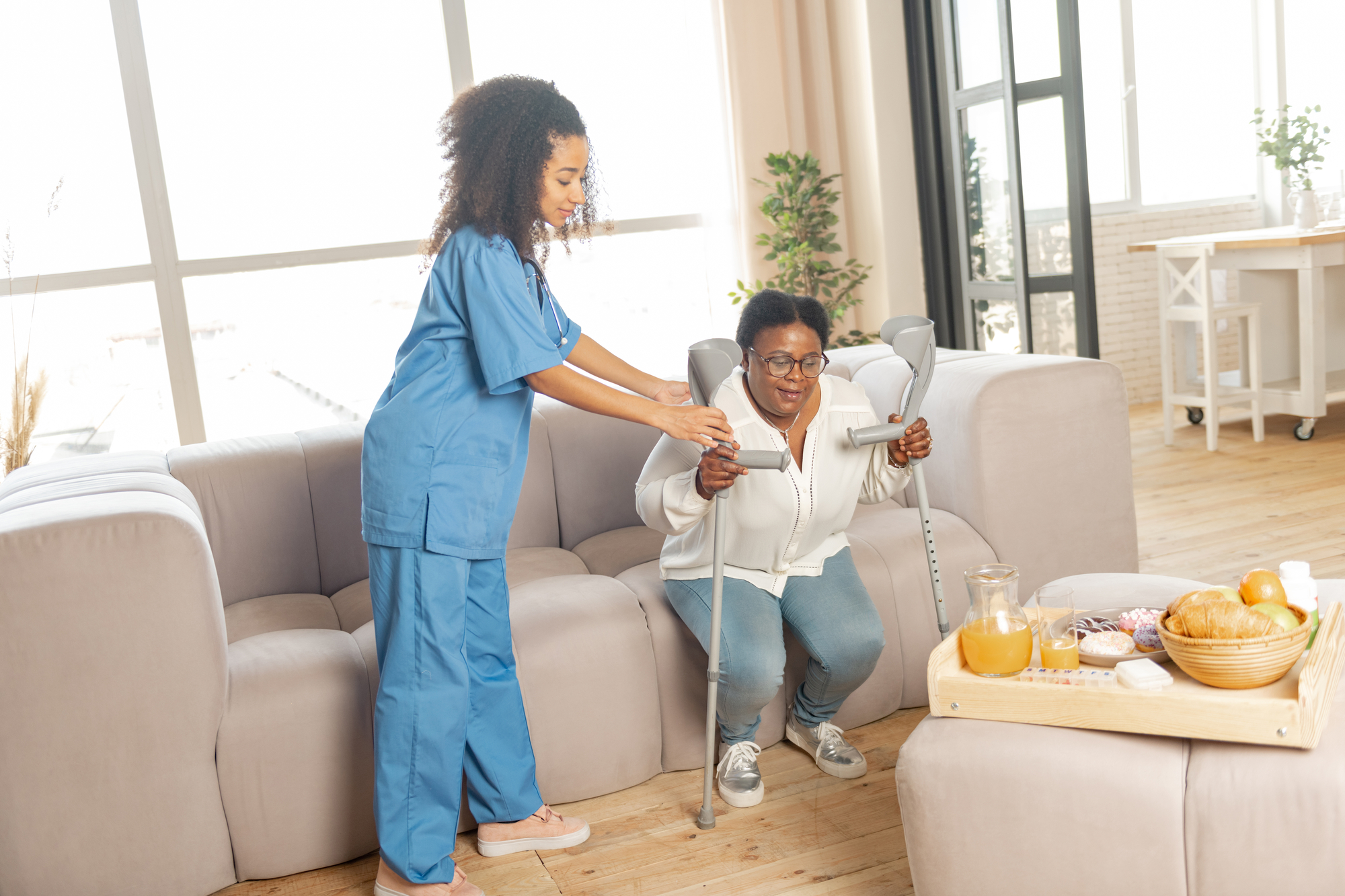 Nurse helping woman to stand from the sofa with crutches